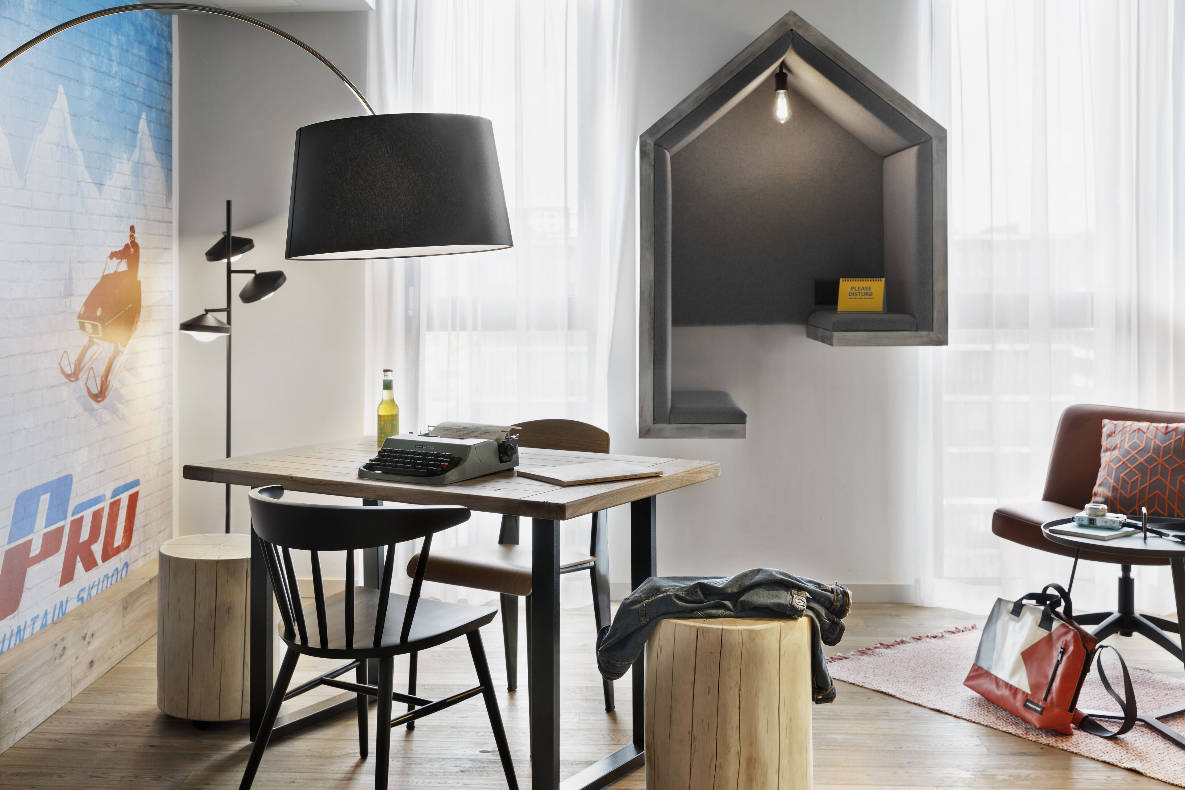 Una habitación moderna con una mesa de madera, una silla negra, una lámpara, un estante pequeño y un sofá.