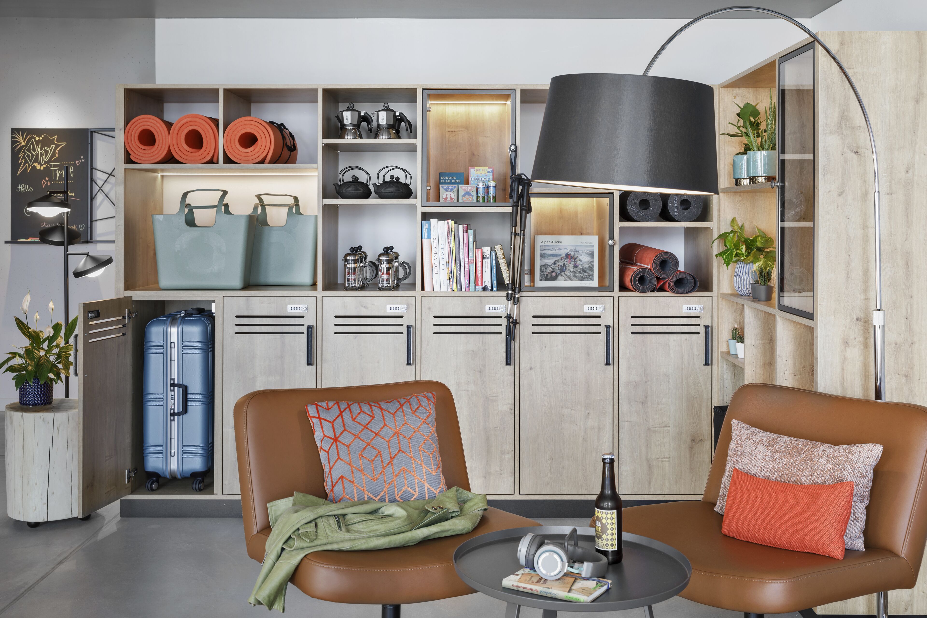 A modern living room with a sofa, a coffee table, and a lamp. There are shelves with books, a suitcase, and decorative items.