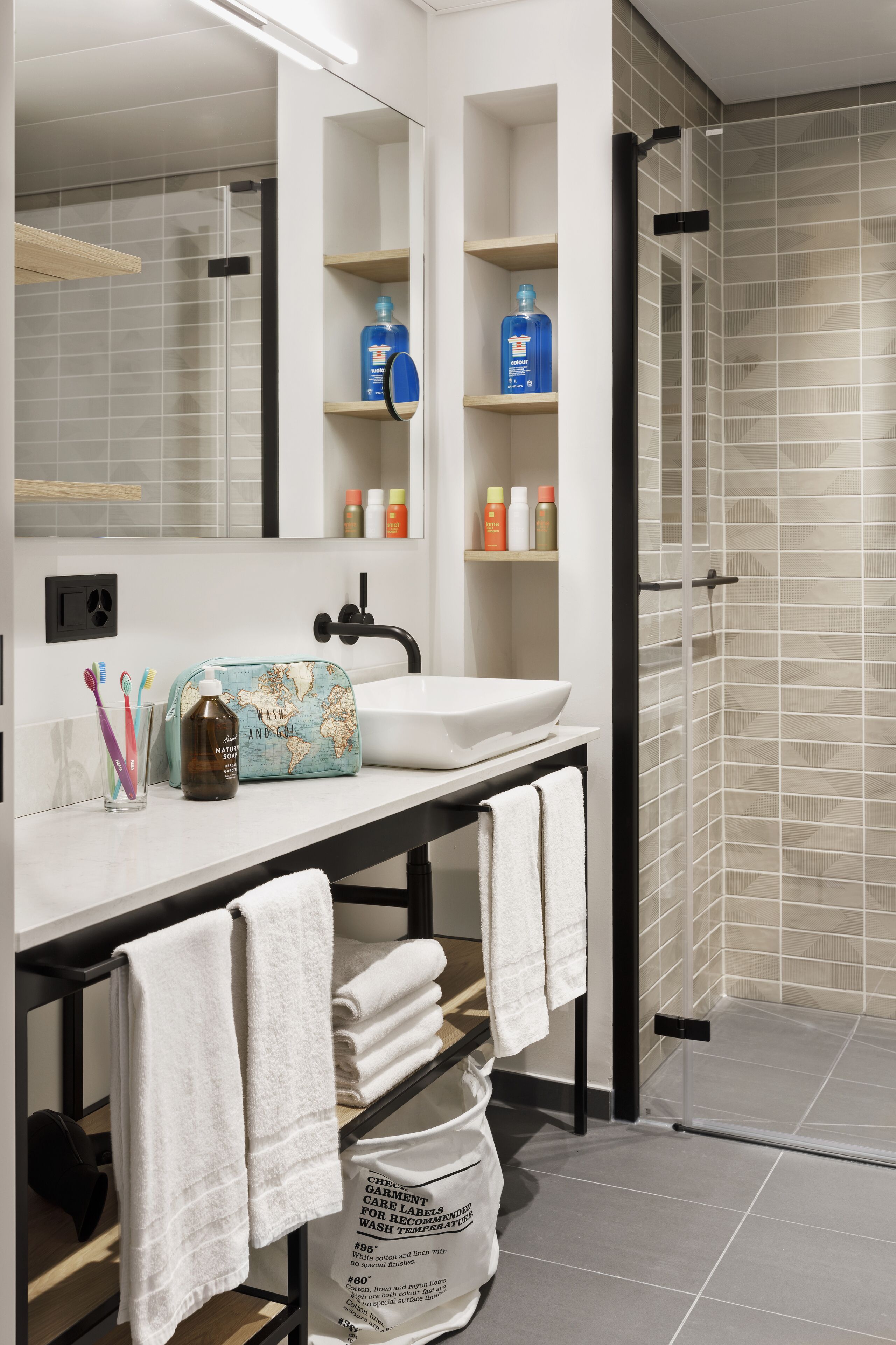 A modern bathroom with a white sink, mirror, and shelves with toiletries. There is a towel rack with towels, a glass shower enclosure, and a map-themed toiletry bag on the counter.
