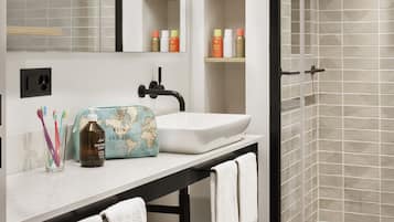 A modern bathroom with a white sink, mirror, and shelves with toiletries. There is a towel rack with towels, a glass shower enclosure, and a map-themed toiletry bag on the counter.