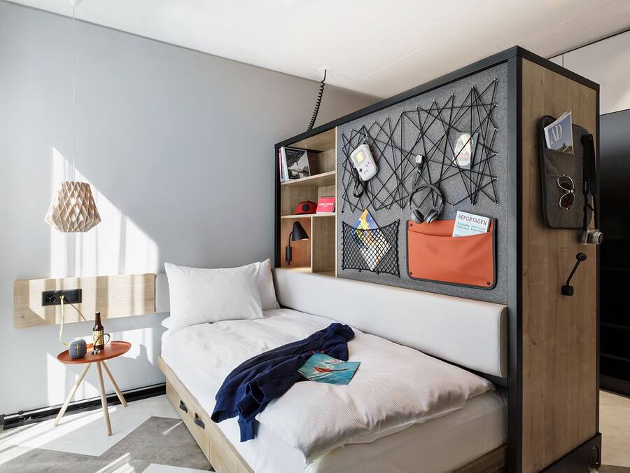Ett modernt hotellrum med en säng, ett litet sidobord, en väggmonterad förvaringsenhet och en hängande lampa.