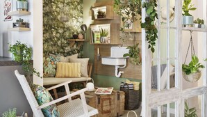 Un salon chaleureux avec un fauteuil en osier blanc, un banc en bois et un canapé agrémenté de coussins à rayures. On y trouve des plantes en pot, un évier blanc et un meuble en bois avec des étagères.