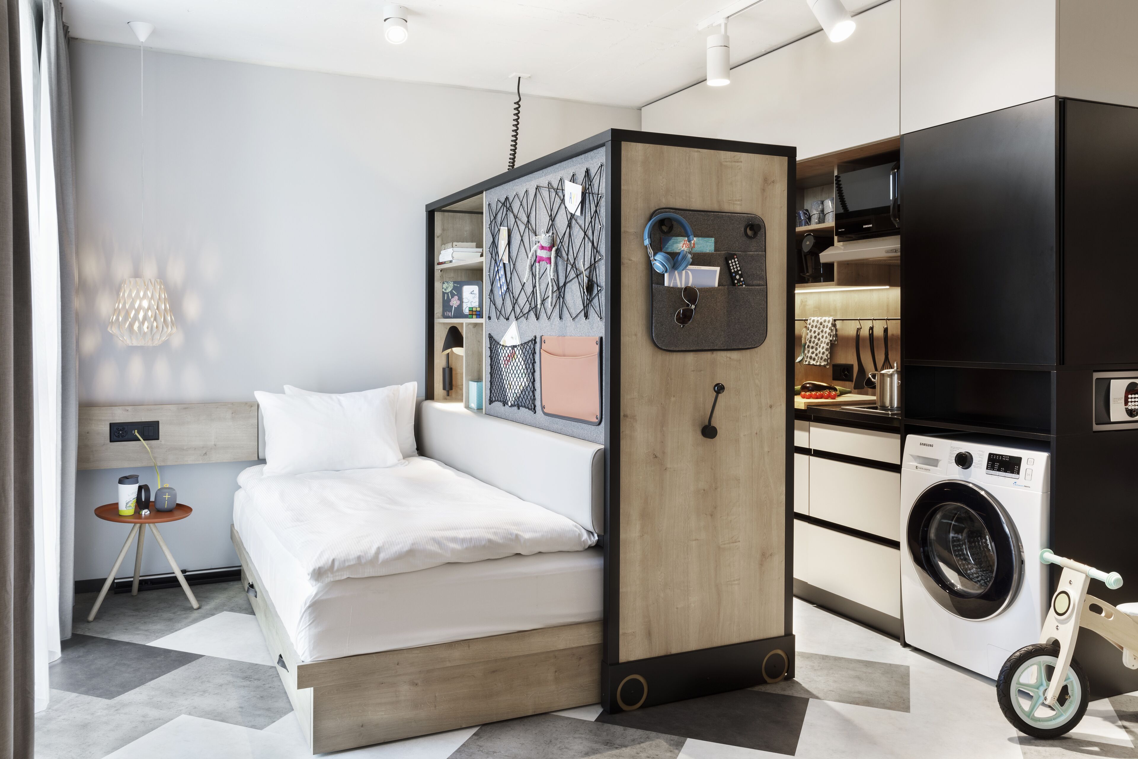 A compact hotel room with a bed, a small table, a washing machine, and a bike.