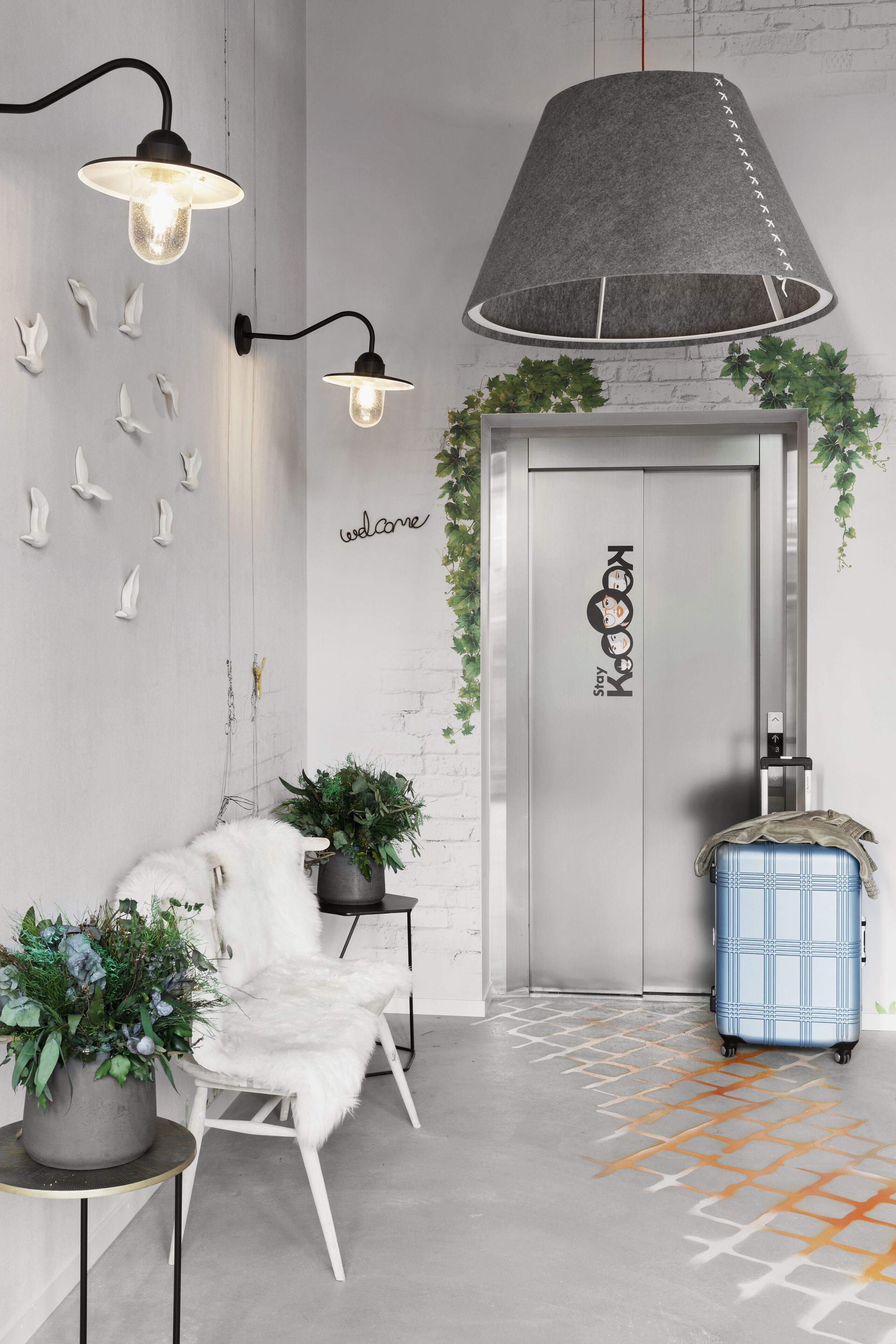 Un hall d’hôtel moderne avec un fauteuil blanc, une valise bleue et un grand luminaire suspendu gris.