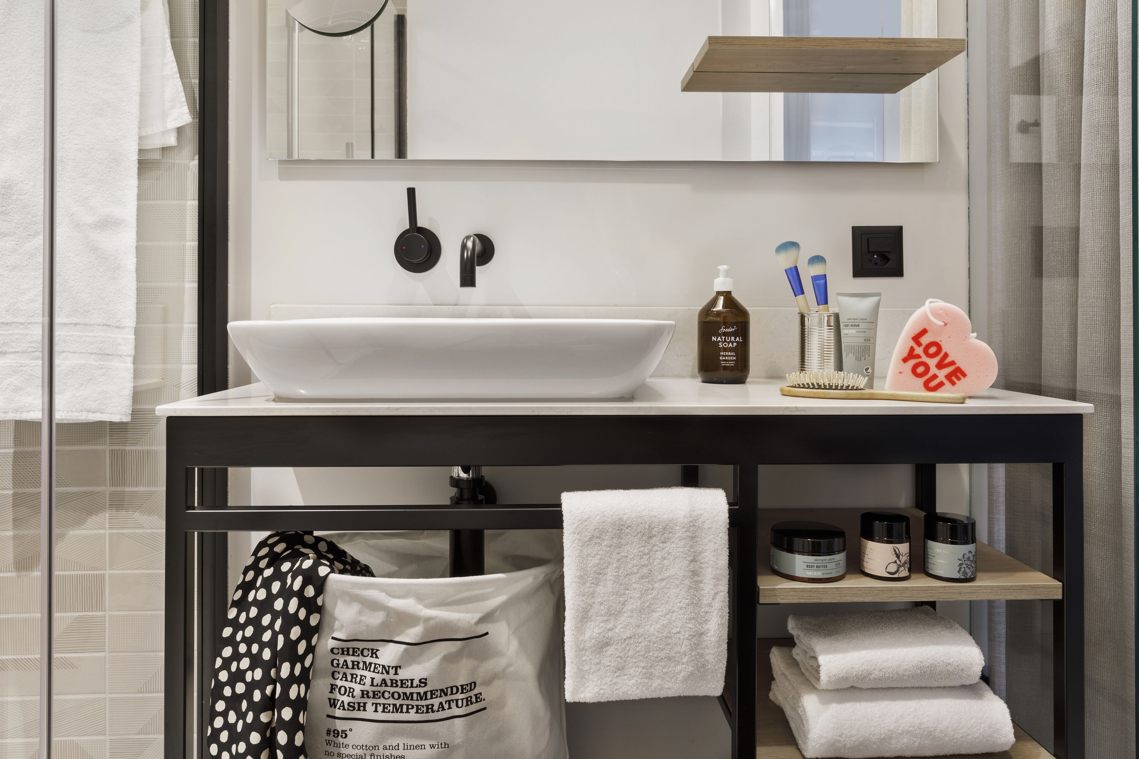 A modern bathroom with a white sink, black faucet, and a mirror above. There are towels, a bag, and toiletries on the counter.