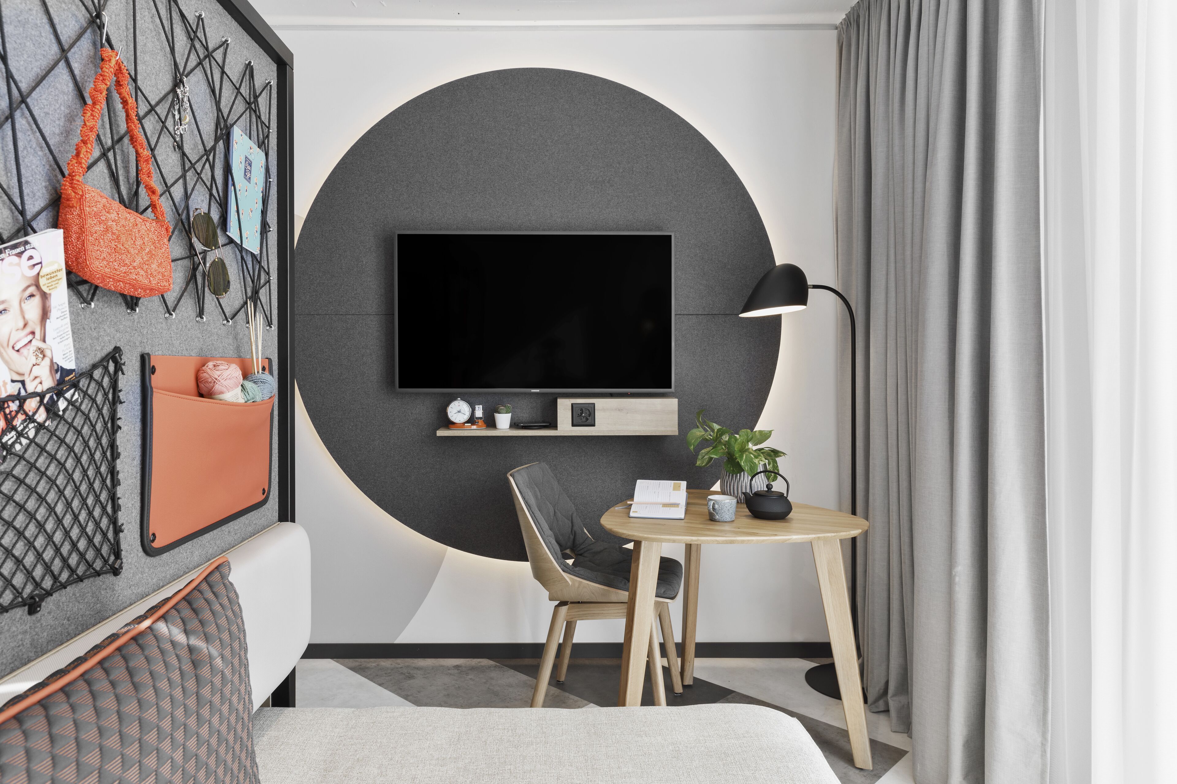 A modern room with a large TV mounted on a circular accent wall, a wooden table with a chair, a potted plant, and a wall-mounted shelf with decorative items.