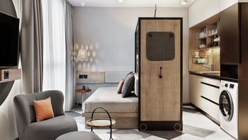 A compact living space with a sofa, a small round table, a bed, a wooden wardrobe, a washing machine, and a TV.
