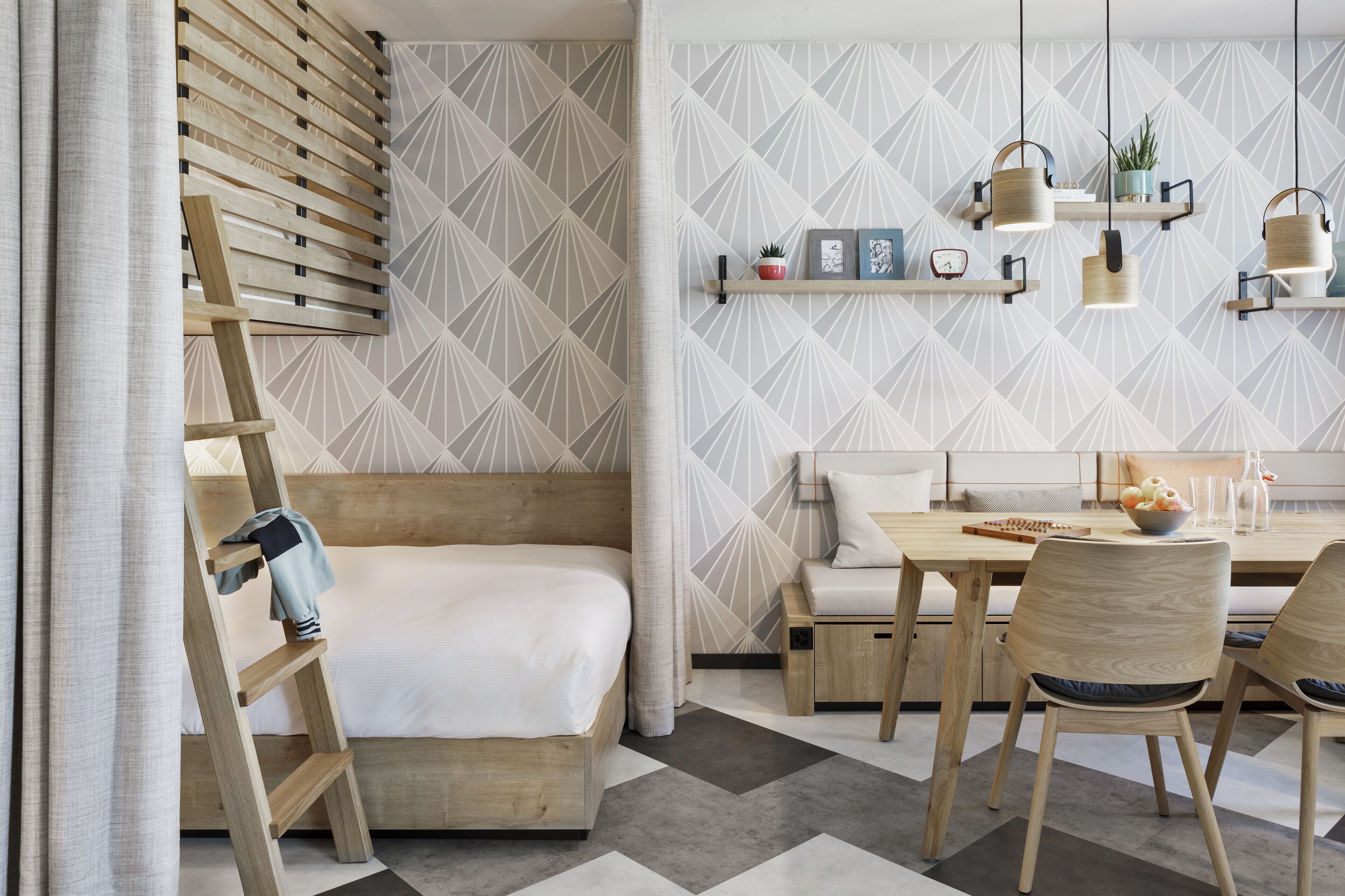 Une chambre moderne avec un lit en mezzanine, un coin repas comprenant une table et des chaises, et un mur recouvert d’un papier peint à motifs géométriques.