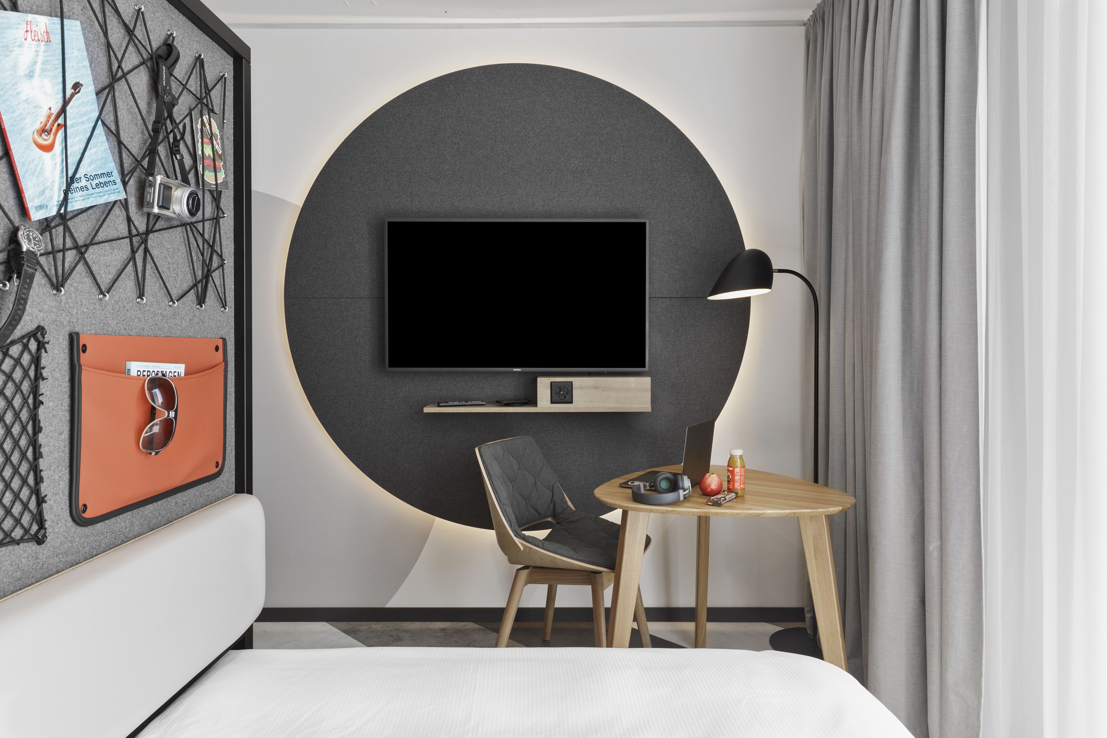 Ett modernt hotellrum med en stor, rund tv som är monterad på väggen, ett litet träbord med en stol och en säng med en vit sänggavel.