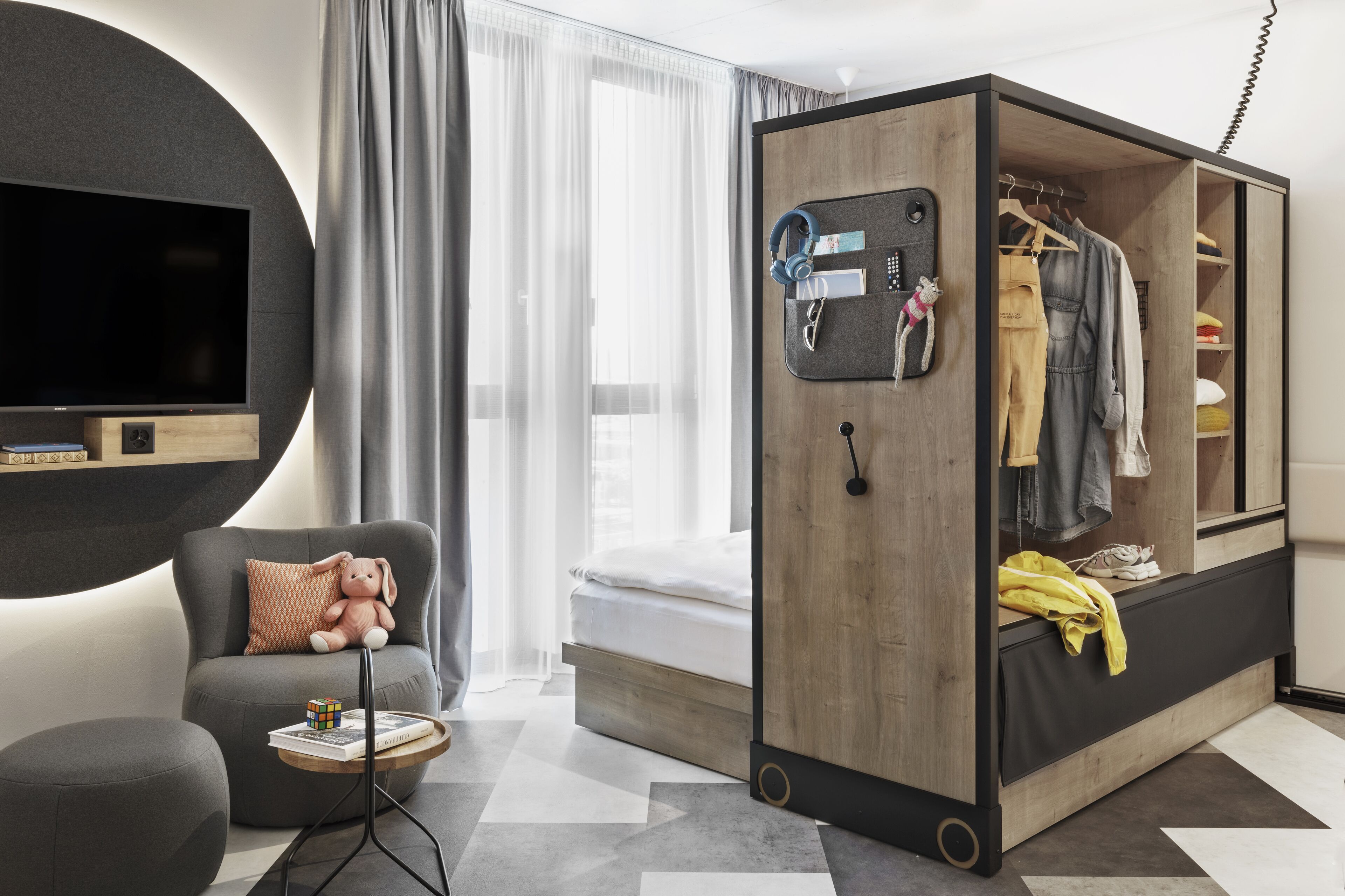 Een moderne hotelkamer met een bed, een zithoek met een comfortabele fauteuil, een kleine tafel met een Rubik's Cube en een kledingkast met daarin hangende kleding.