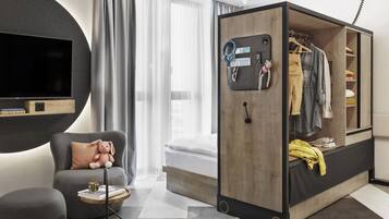 Een moderne hotelkamer met een bed, een zithoek met een comfortabele fauteuil, een kleine tafel met een Rubik's Cube en een kledingkast met daarin hangende kleding.