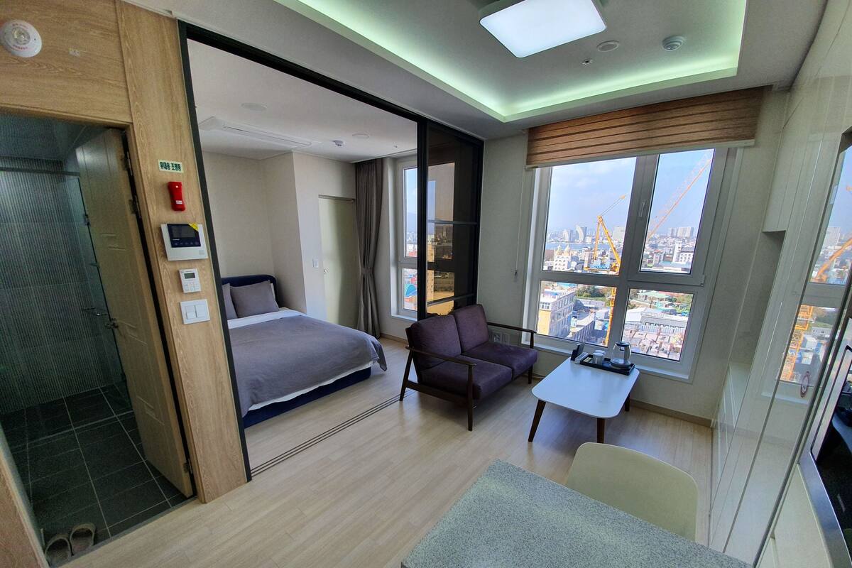 A modern hotel room with a bed, a sofa, a dining table, and a view of the city.