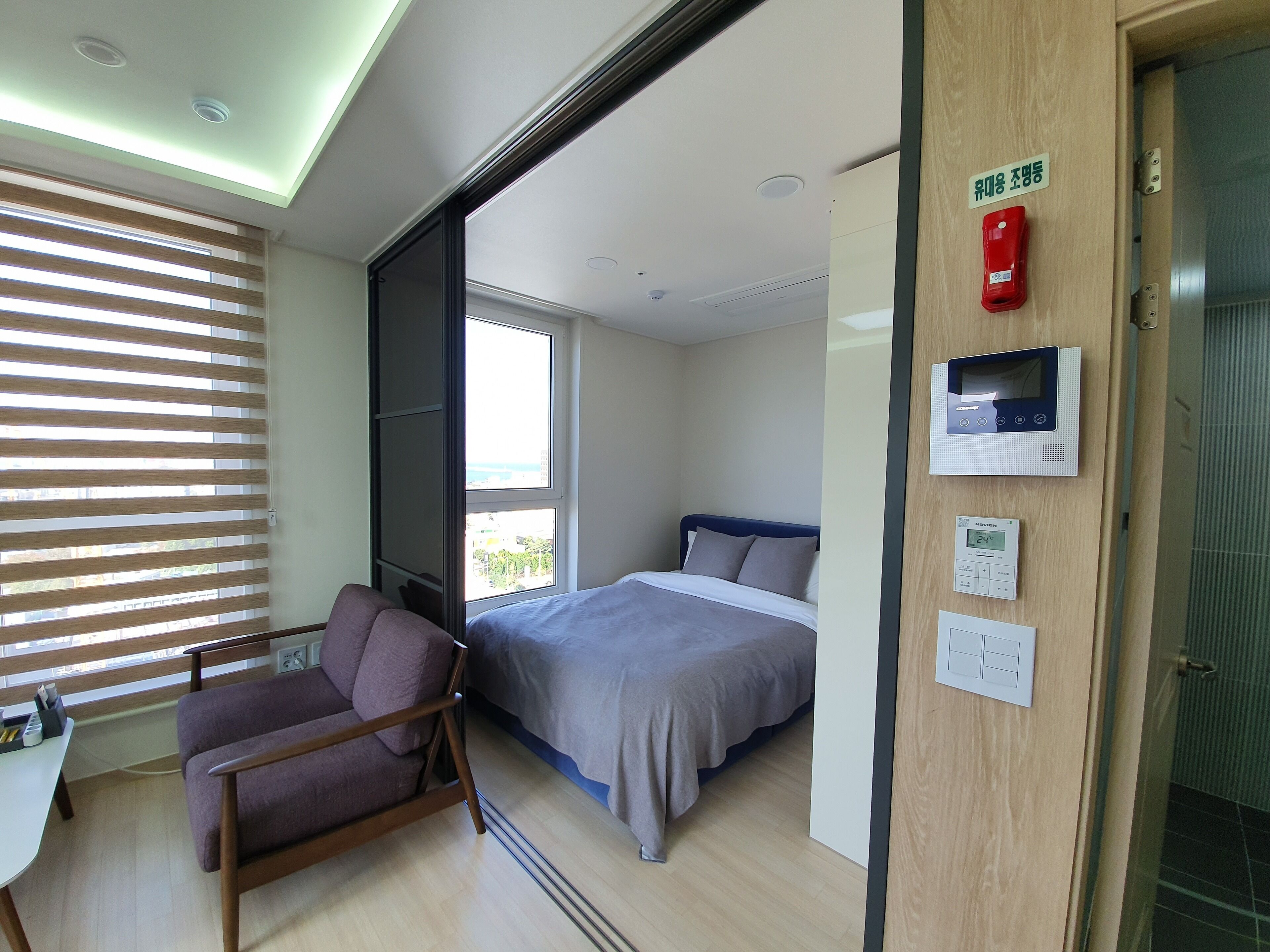 A hotel room with a bed, a chair, a desk, a window with blinds, a thermostat, and a fire alarm.