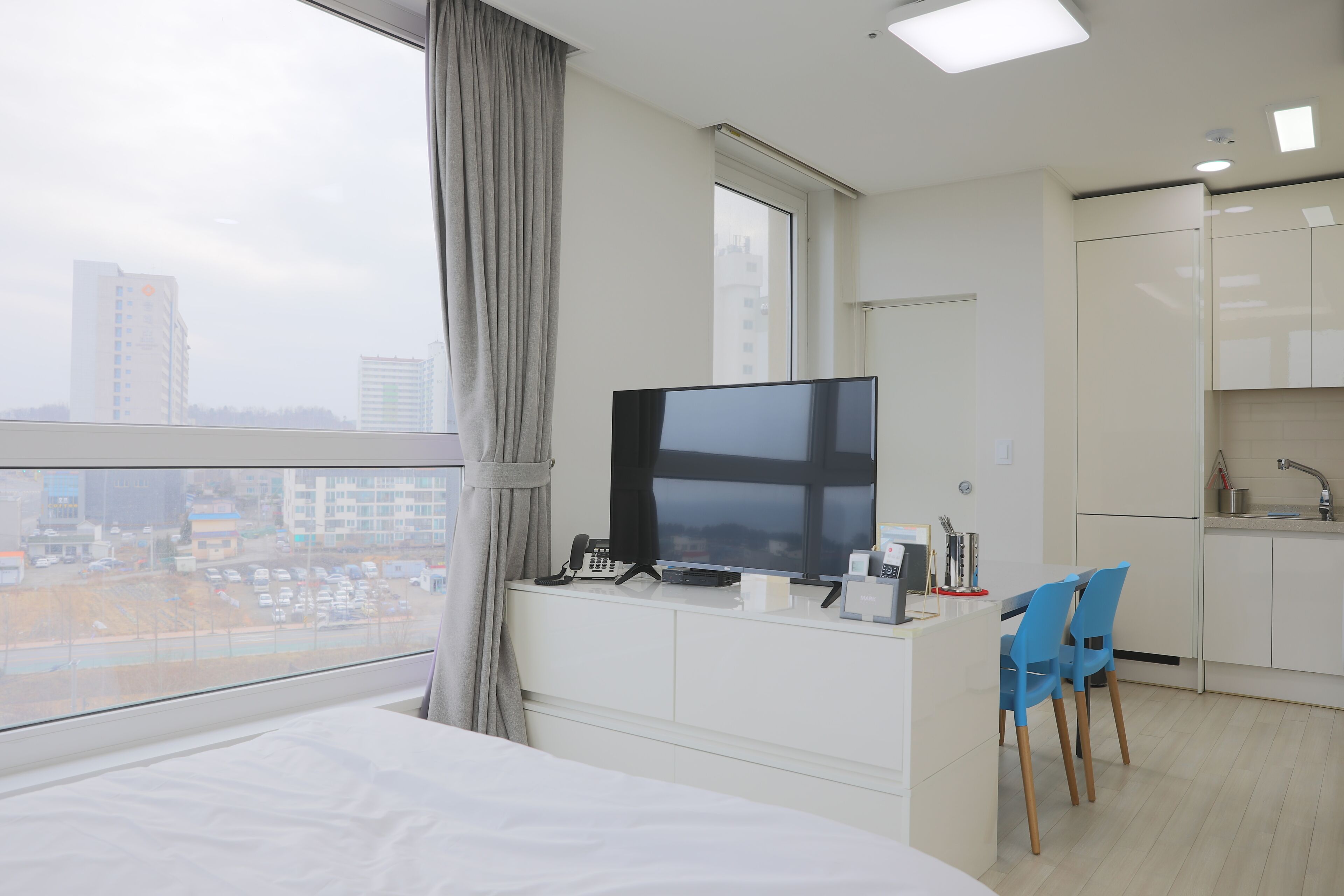 A modern hotel room with a large bed, a TV on a desk, a view of the city, and a kitchenette.