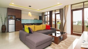 A modern living room with a grey sectional sofa, a coffee table, and a dining area with a white table and lamp. There is a kitchen in the background with a fridge and cabinets. - Rozelle Hotel (Bandung)