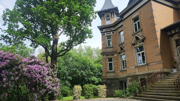 A large brick building with a tower, surrounded by lush greenery and flowering bushes. - Hotel Am Park (Reichenbach im Vogtland)