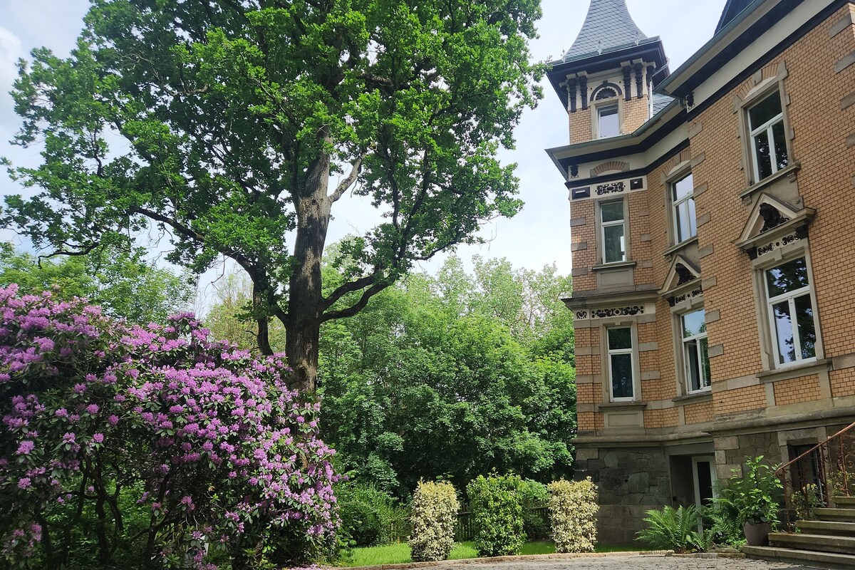 A large brick building with a tower, surrounded by lush greenery and flowering bushes.
