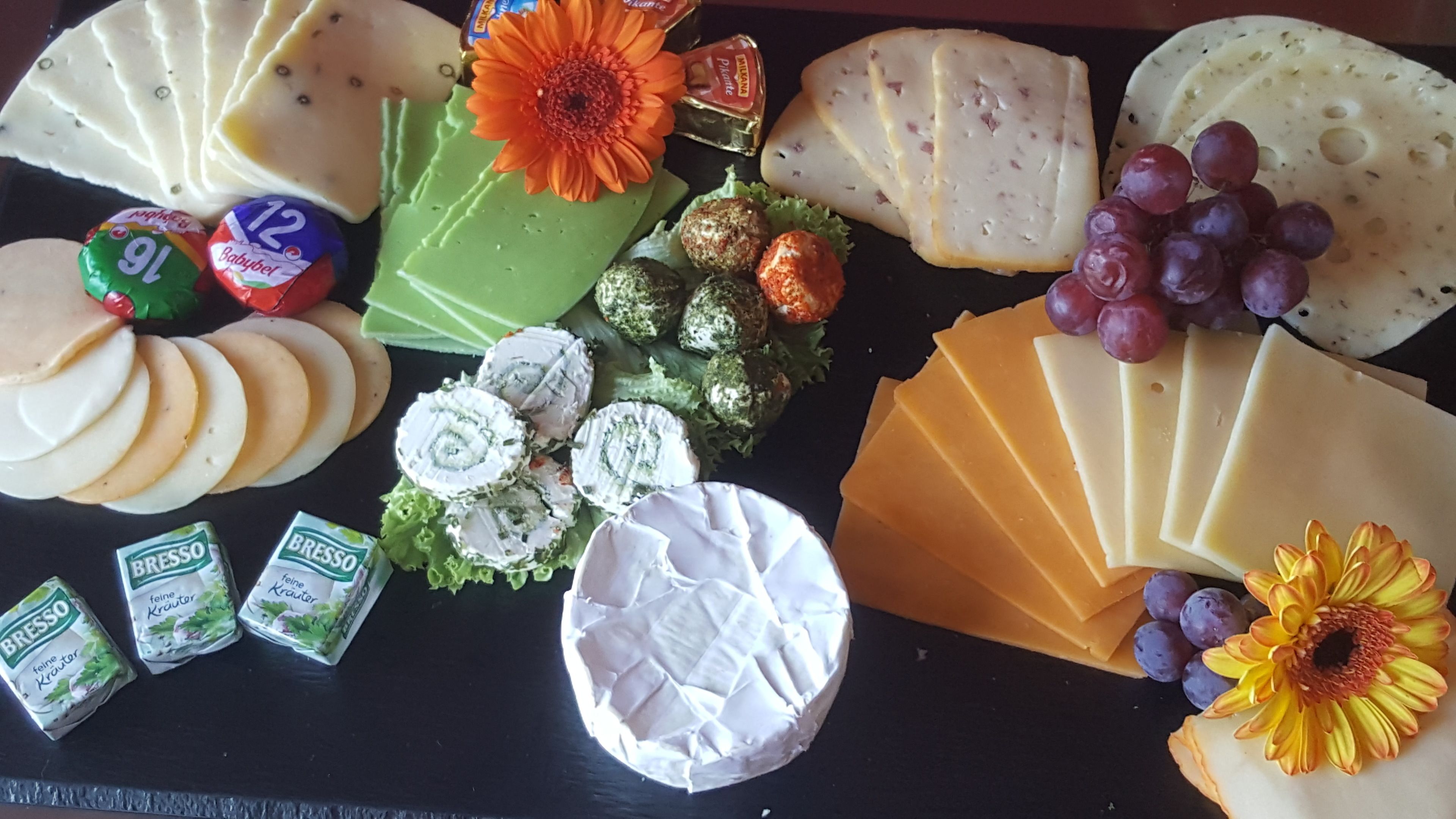 A variety of cheeses, including Brie, Gouda, and Camembert, arranged on a black table with decorative flowers and grapes.
