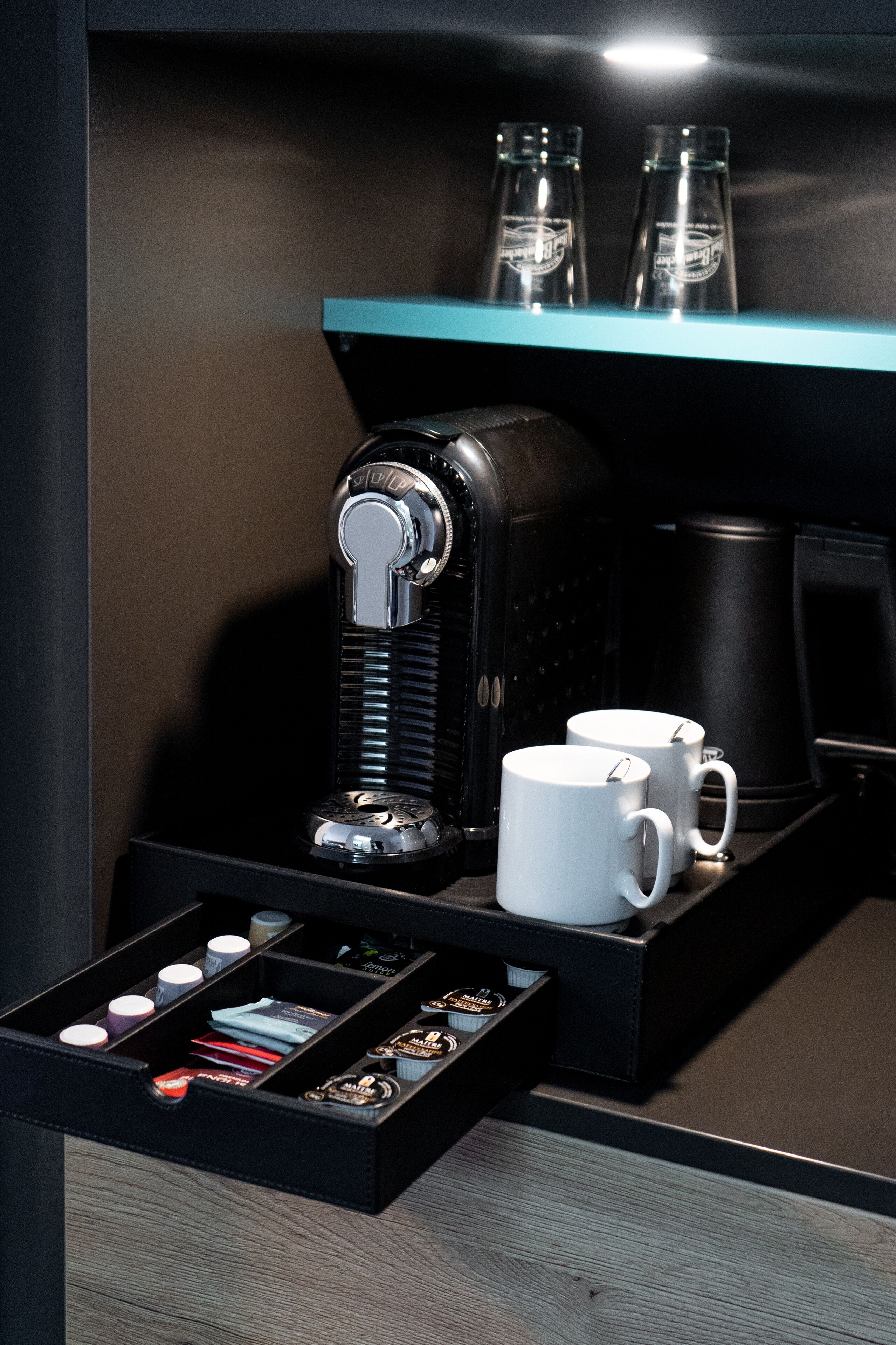 Une machine à café avec une verseuse, deux tasses et un tiroir contenant des dosettes de café et une cuillère.