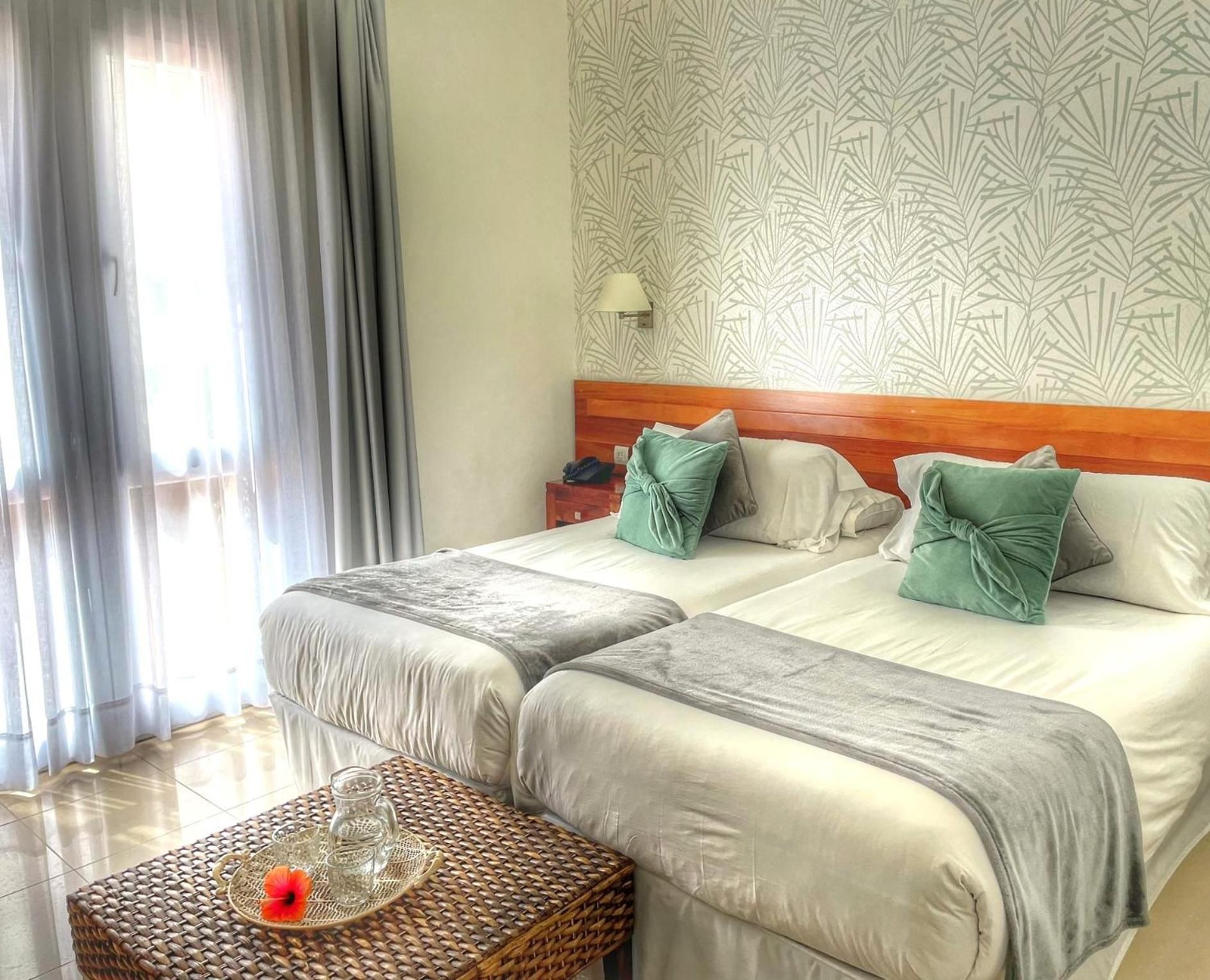 A hotel room with two beds, a wooden headboard, a bedside table with a lamp, and a woven table with a glass pitcher and flower.