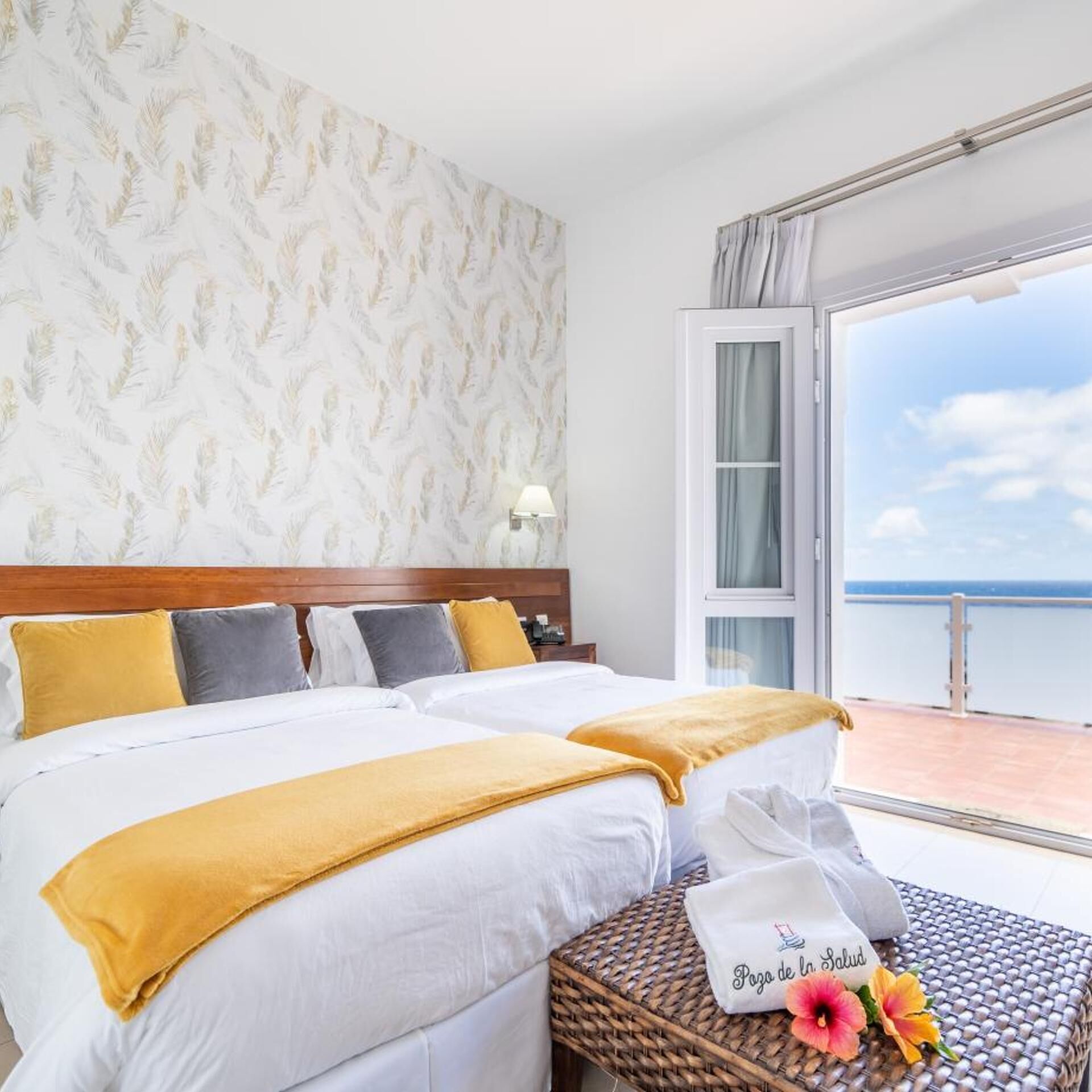 Habitación de hotel con dos camas, un balcón con vista al mar, papel tapiz estampado y una mesita de noche con una toalla y un almohadón.