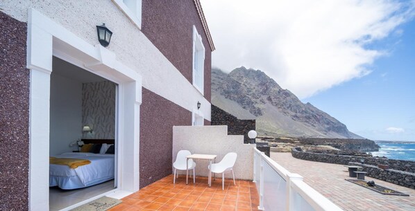 Quarto com cama e mesa de cabeceira, varanda com mesa e cadeiras, e vista para o oceano e montanhas.