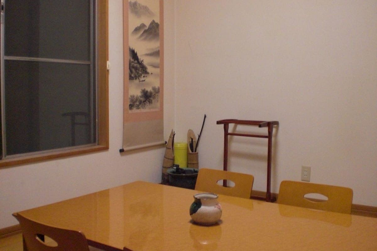 A dining area with a wooden table, two chairs, a vase, and a painting on the wall.