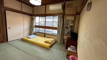 A traditional Japanese room with tatami flooring, a futon bed, a small wooden desk, a pink bucket, and a window with curtains.