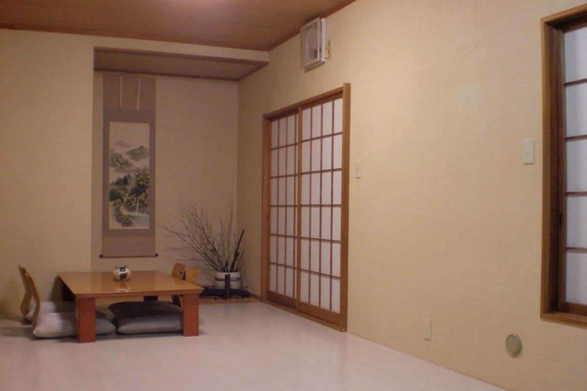 A room with a wooden dining table, chairs, a potted plant, a sliding shoji screen, and a framed artwork on the wall.