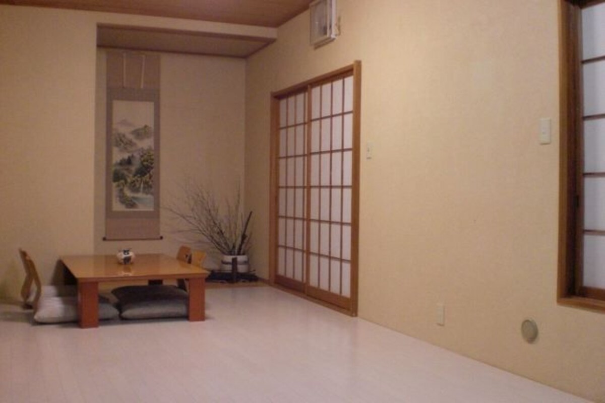 Una habitación japonesa tradicional con suelo de tatami, una mesa de madera baja con cojines, un biombó shoji y un cuadro en la pared.