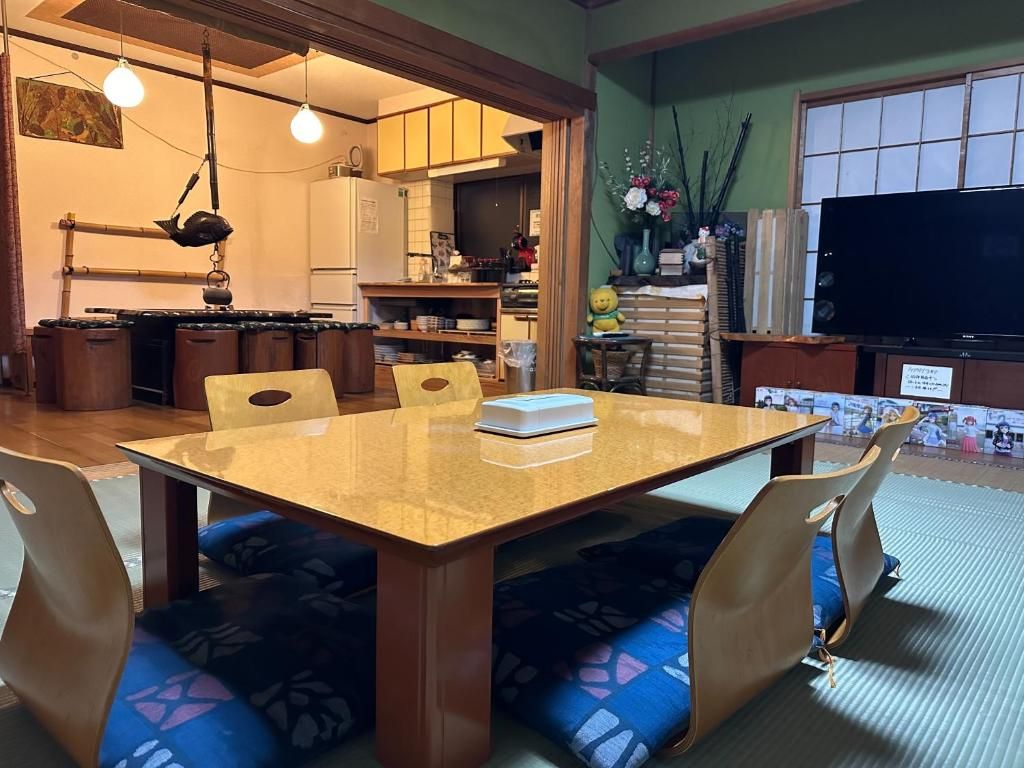 A dining area with a wooden table and chairs, a kitchen in the background, and a television on the right.
