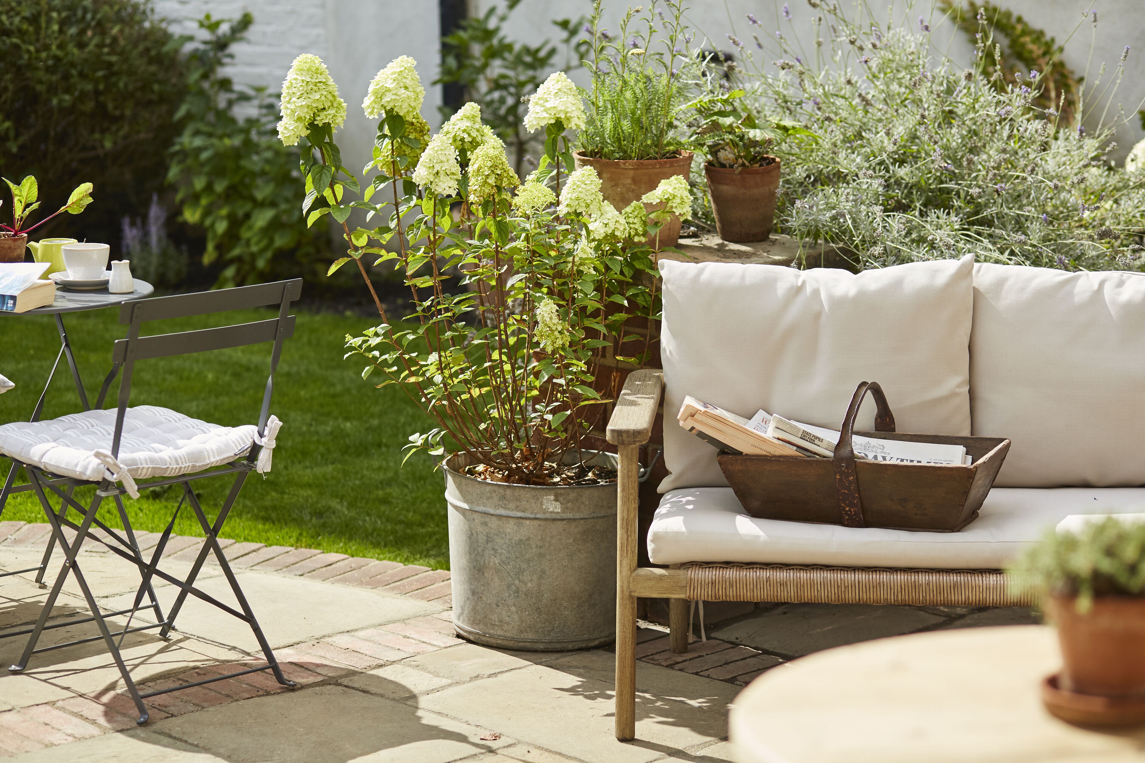 An outdoor seating area with a wicker chair, a cushioned bench, and a table with a teapot and cups.