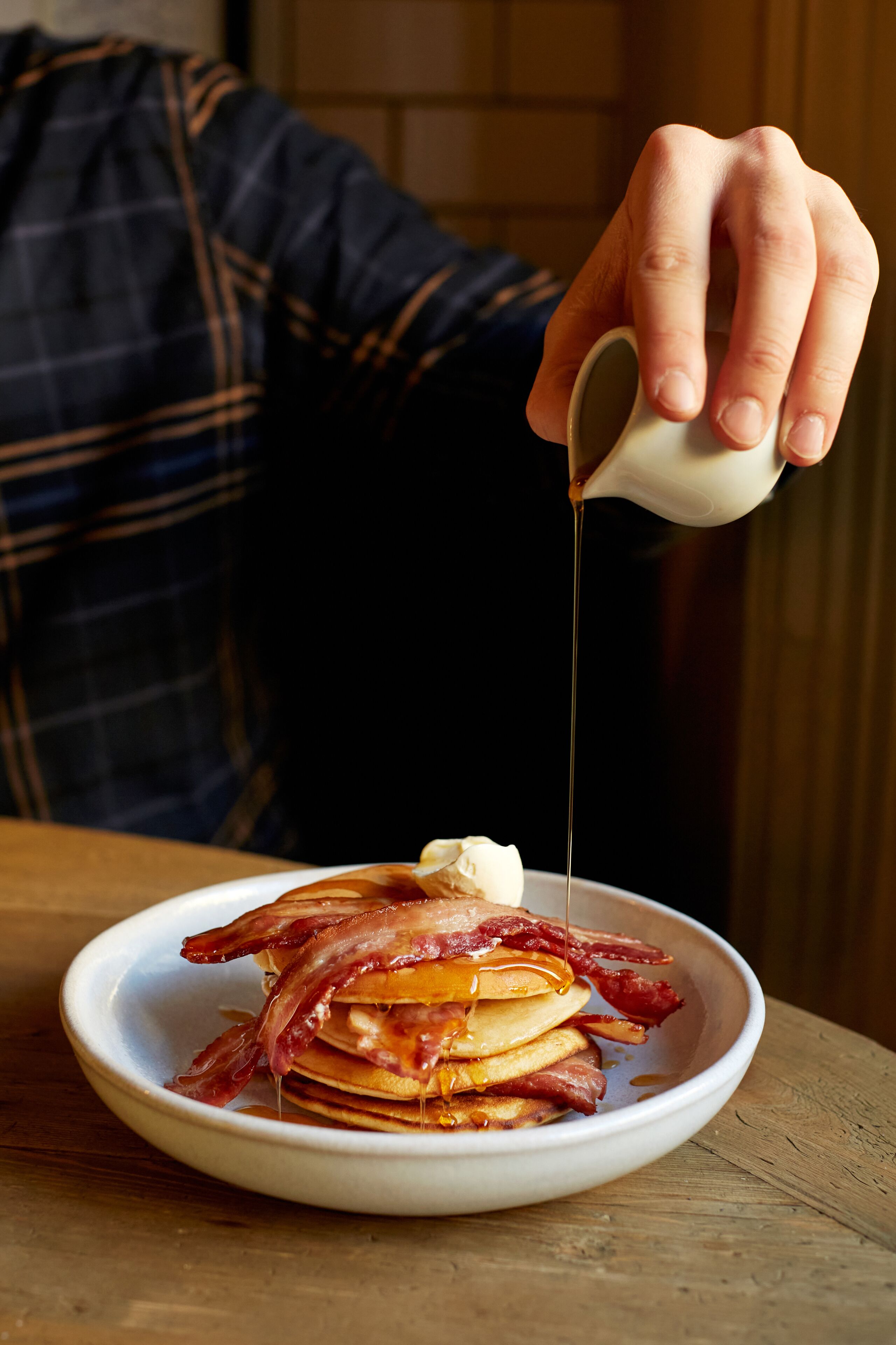 A hand pouring syrup over a stack of pancakes with bacon.