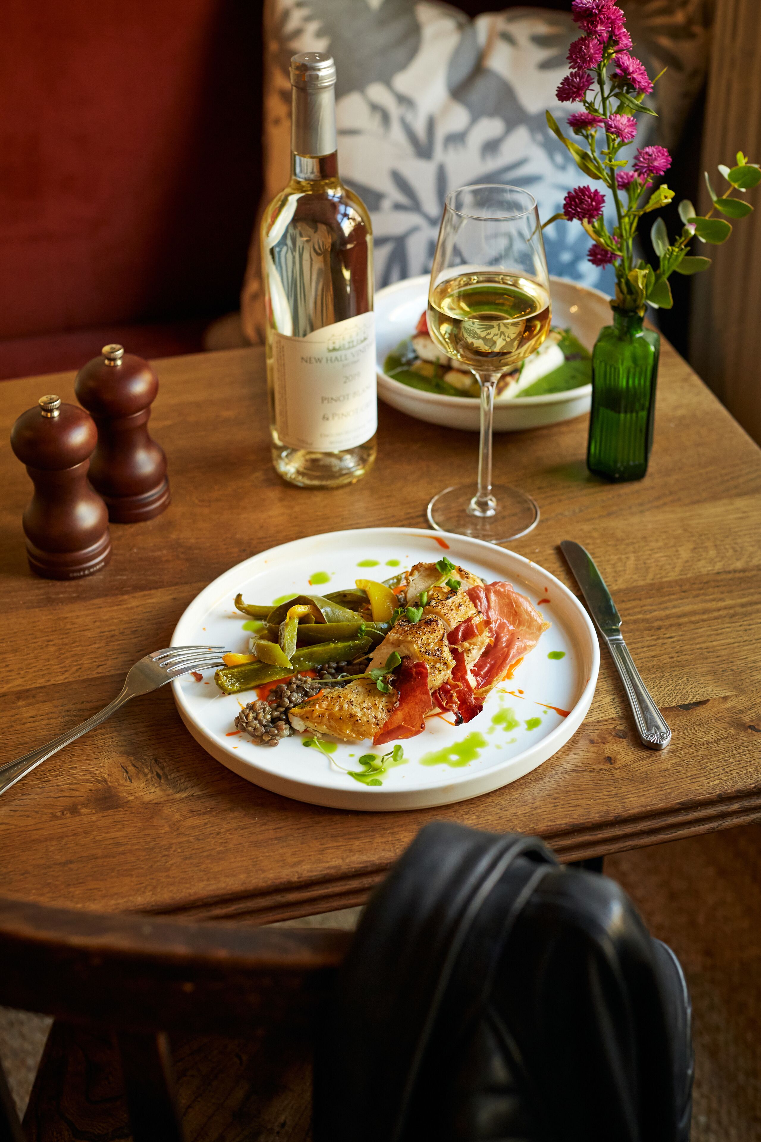 A wooden table with a plate of food, a bottle of wine, a glass of wine, and a vase with flowers.