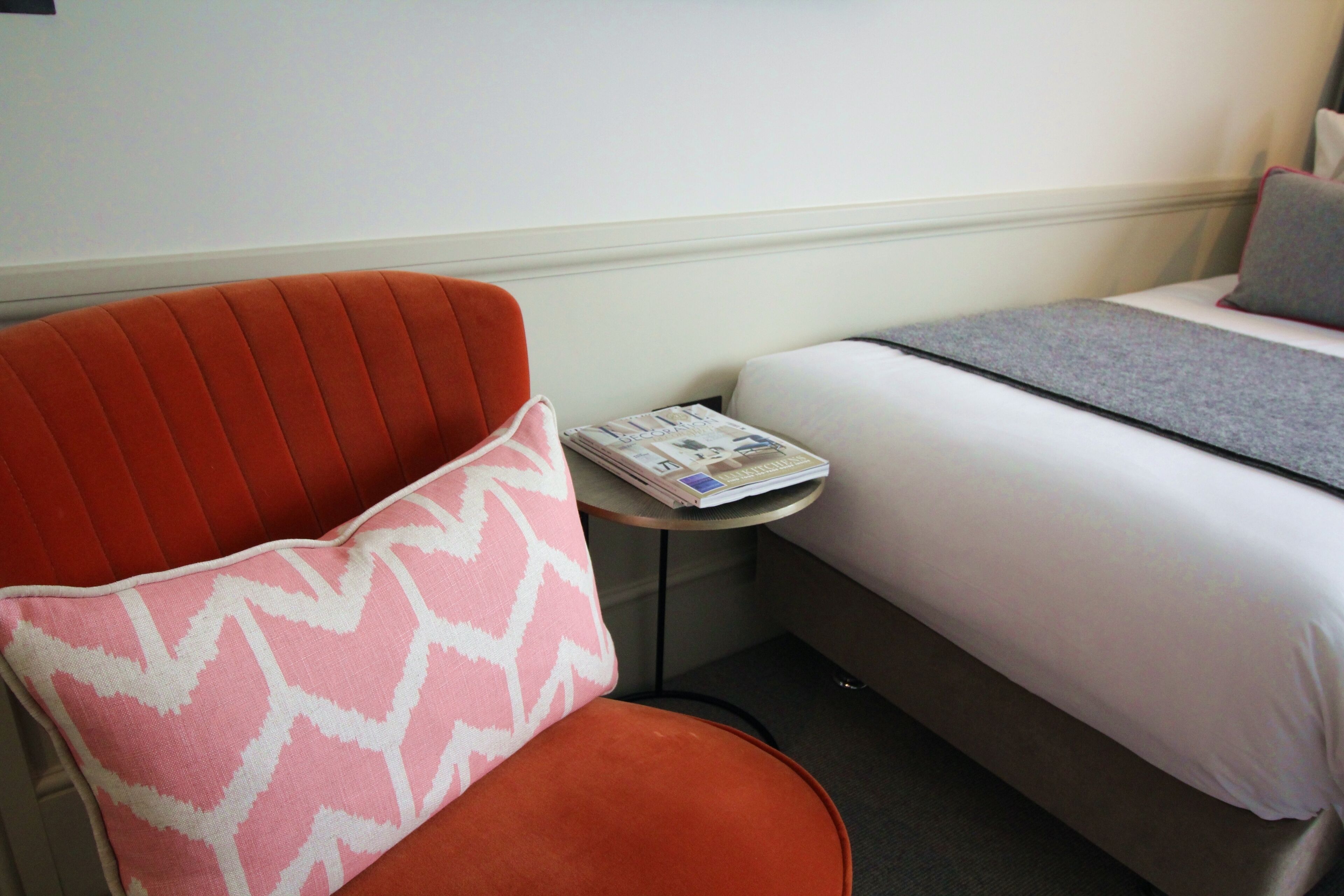 A hotel room with a single bed, a small table with a magazine, and a red armchair with a patterned cushion.