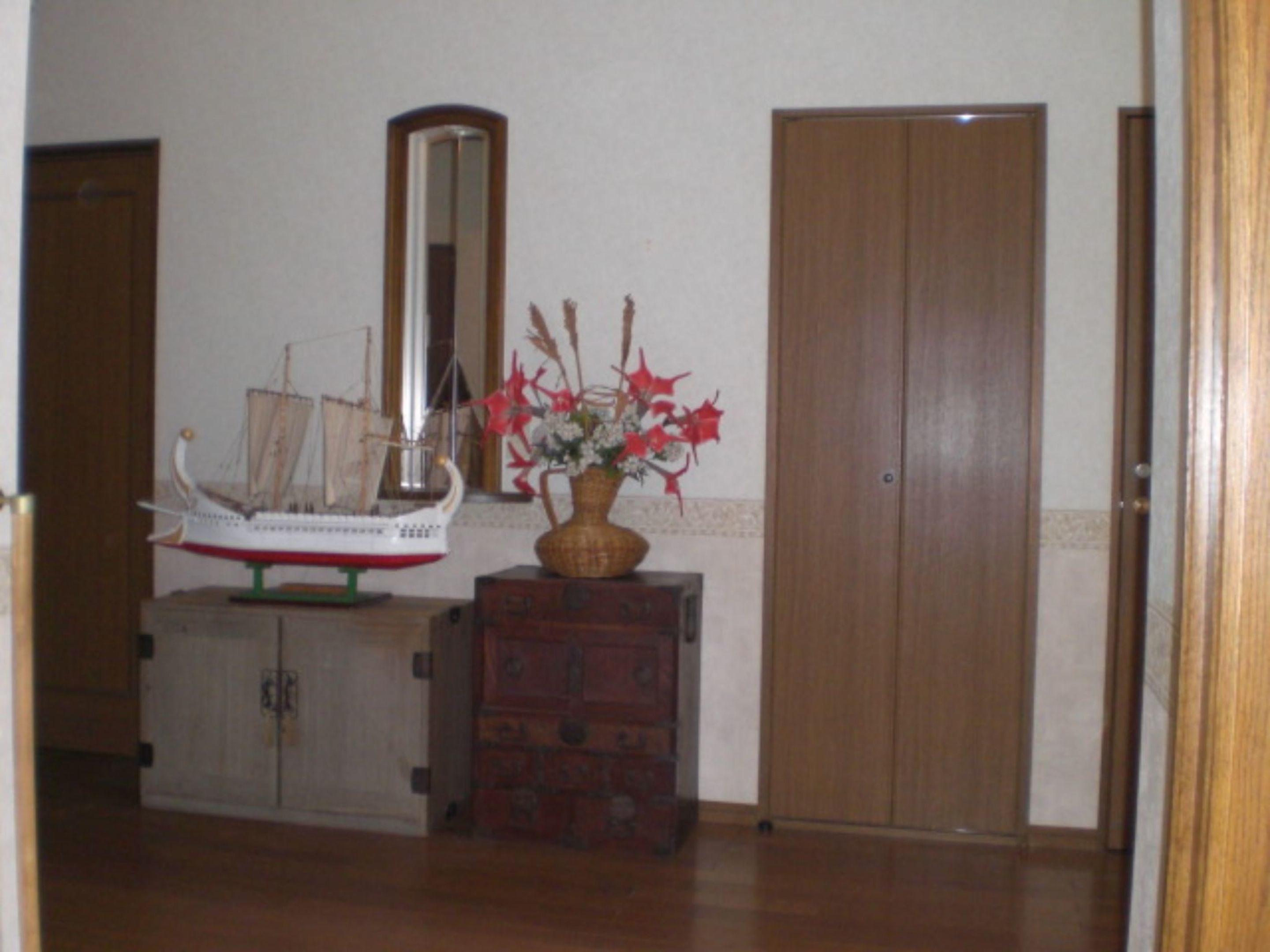 A room with a wooden cabinet, a ship model, a mirror, and a vase with red flowers.