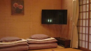 Une pièce de style japonais traditionnel, avec un sol recouvert de tatamis, un lit bas agrémenté de coussins, un téléviseur fixé au mur et un paravent en papier de riz (shoji).