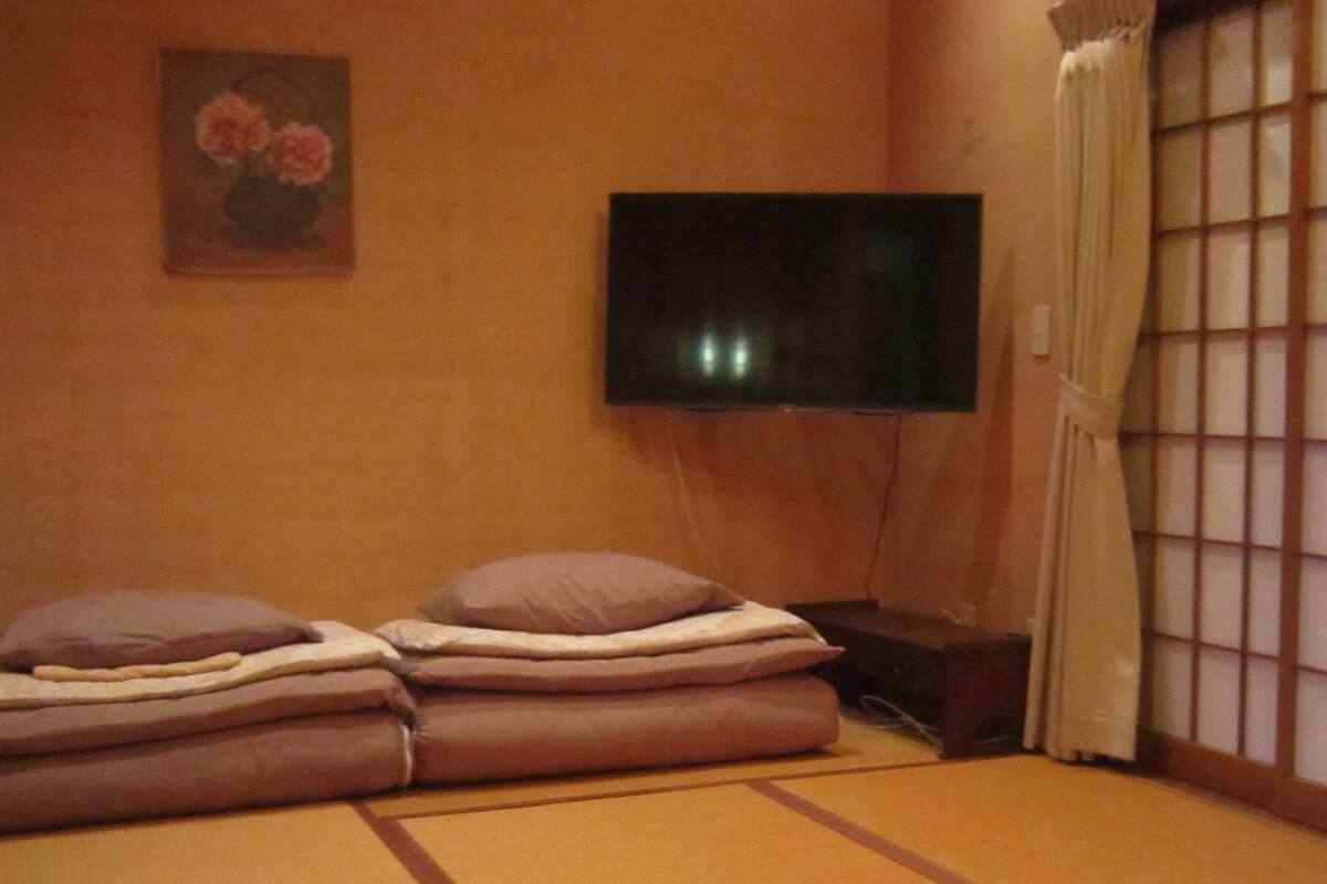 Um quarto tradicional japonês com tatames, uma cama baixa com almofadas, uma televisão na parede e um biombos shoji.