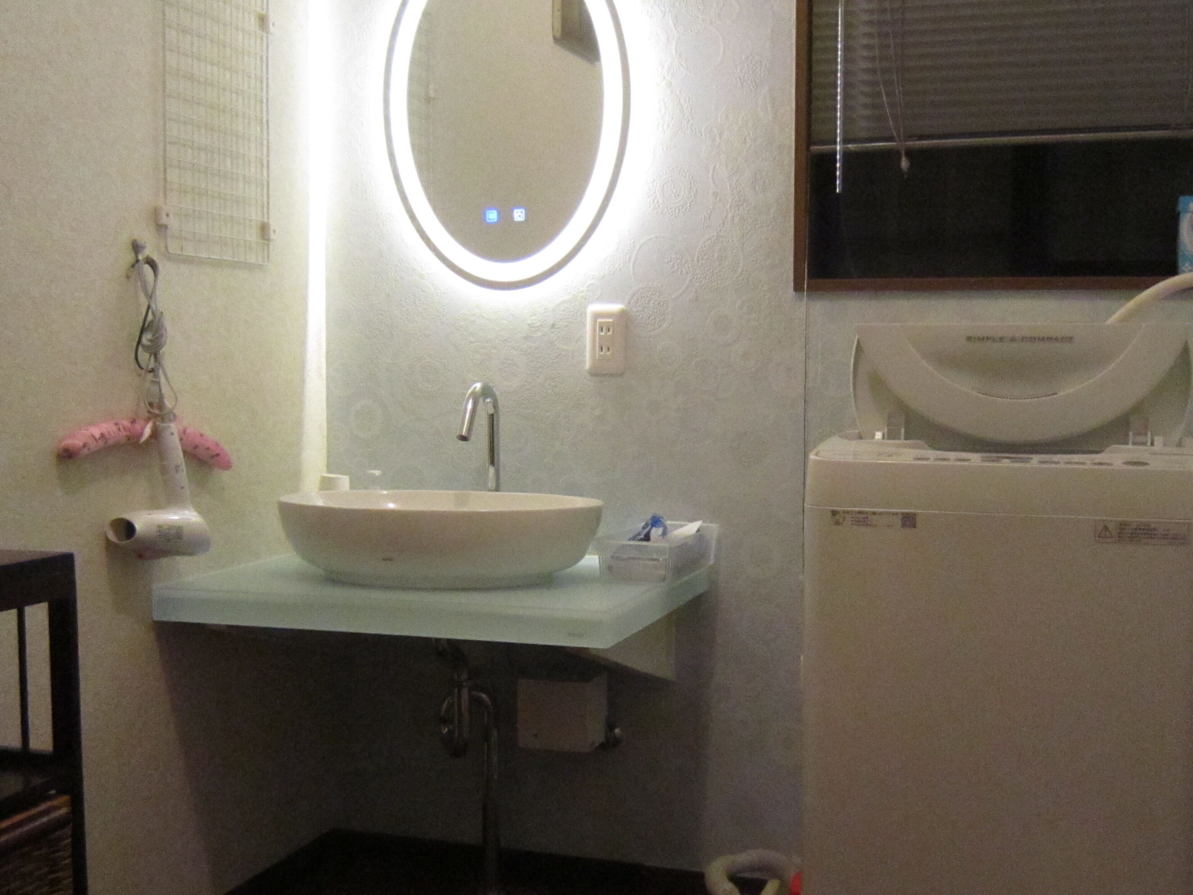 Une salle de bain avec un lavabo blanc, un miroir avec un éclairage intégré, un porte-serviettes et un porte-rouleau de papier toilette.