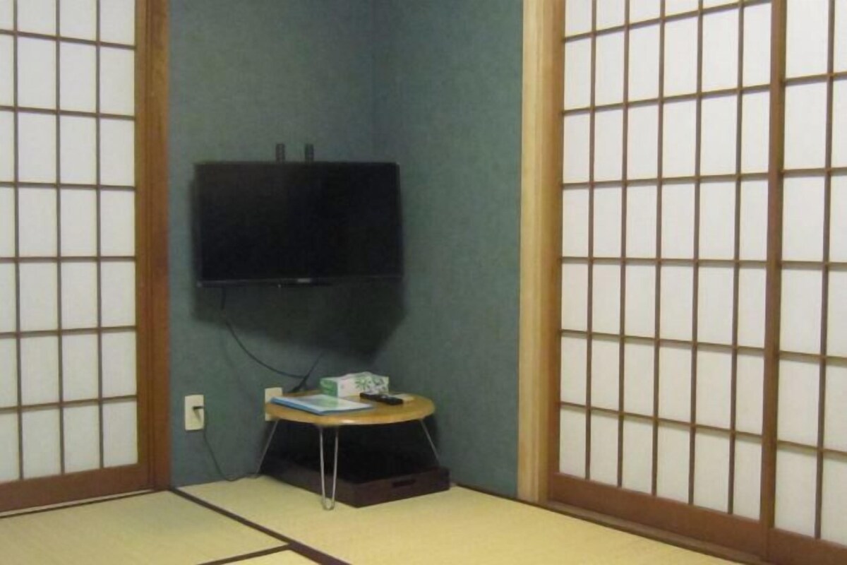 A room with tatami flooring, a television mounted on the wall, a small table with a remote control, and a sliding door with a grid pattern.