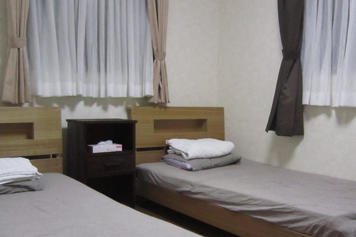 Quarto de hotel com duas camas de solteiro, uma mesinha de cabeceira de madeira com lenços de papel e uma janela com cortinas.