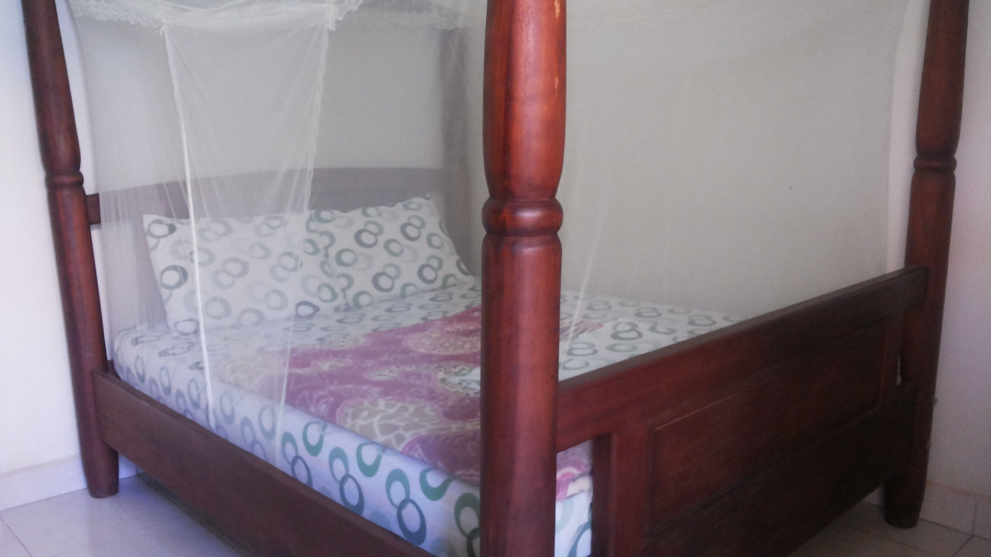 A wooden four-poster bed with a mosquito net, patterned bedding, and a plain wall background.