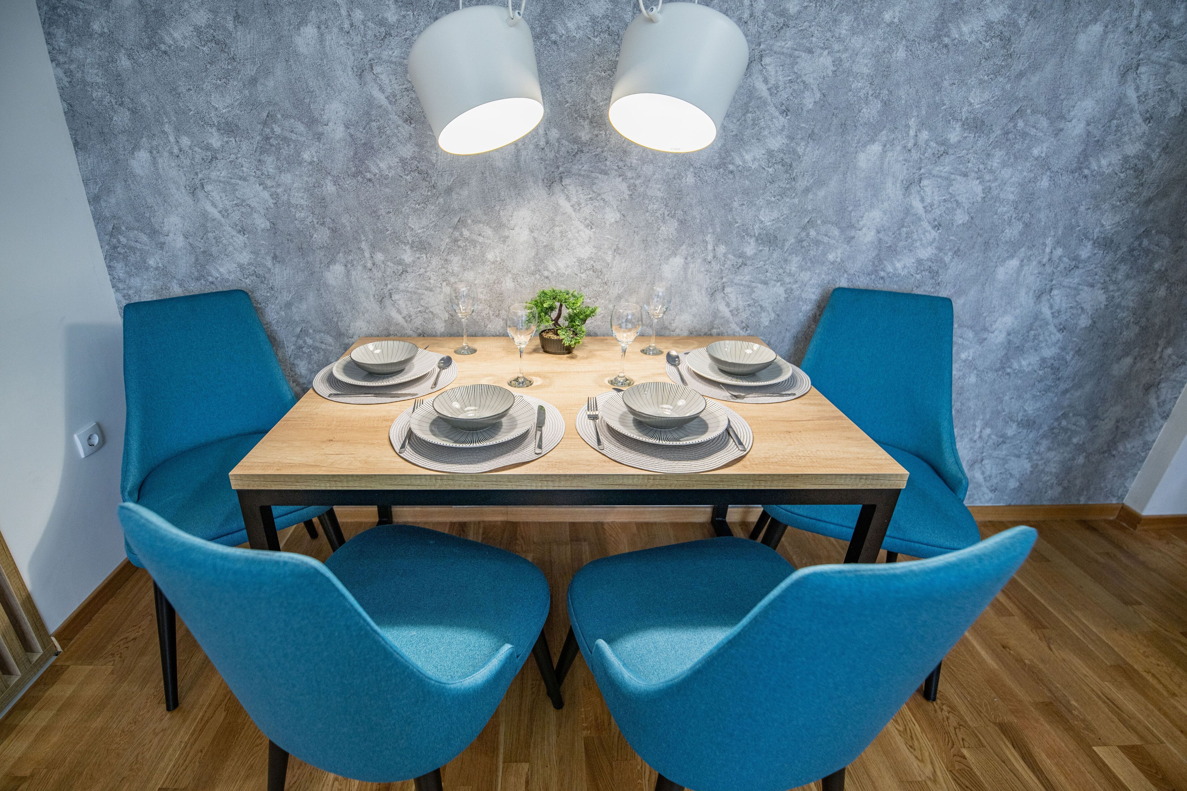 A dining table with blue chairs, two hanging light fixtures, and a small plant centerpiece.