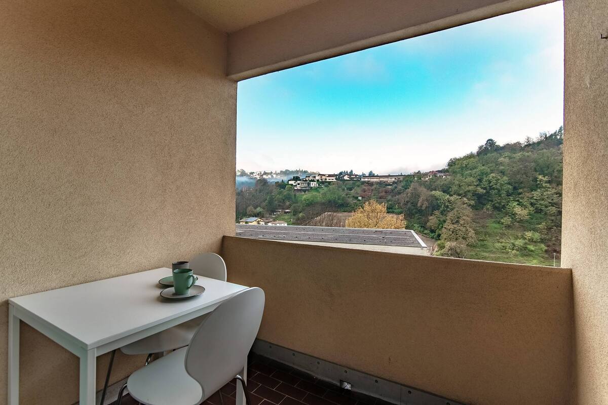 A balcony with a white table and chairs, a green cup, and a view of a hilly landscape with buildings.