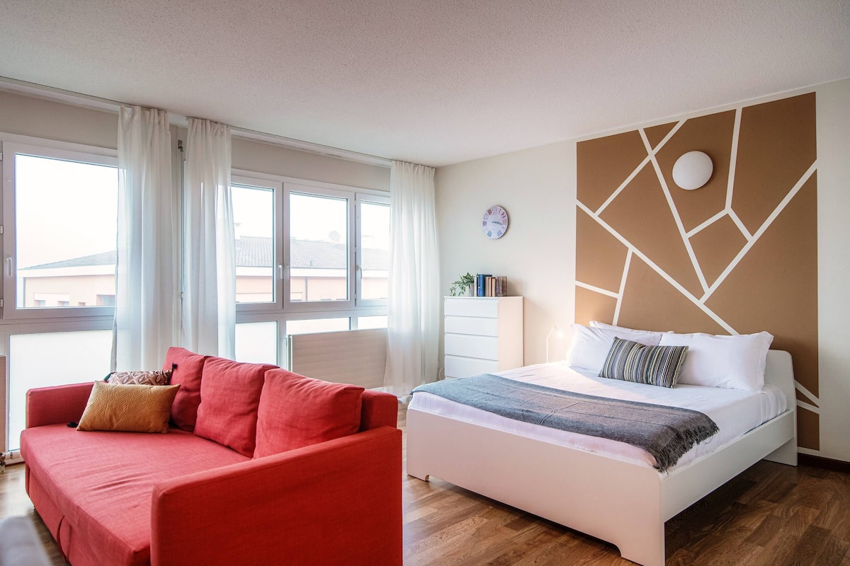 A hotel room with a red sofa, a white bed, a geometric wall design, a clock, and a window with curtains.