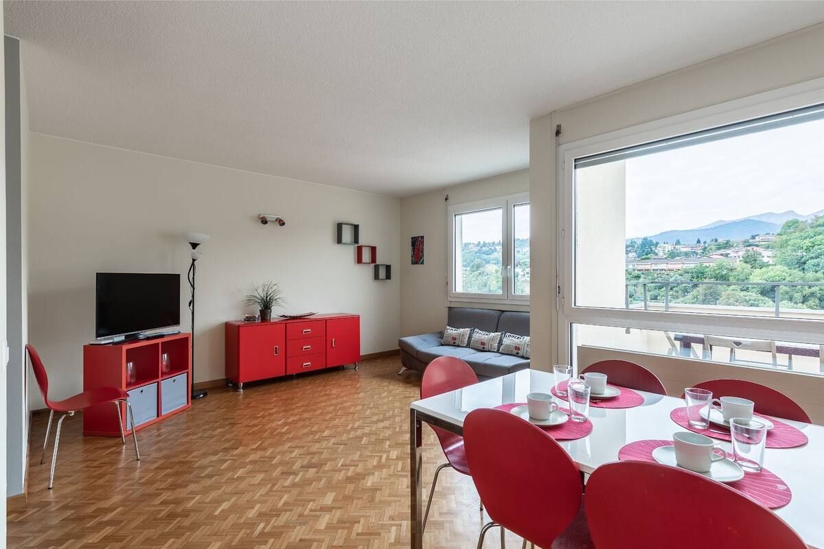 A modern living room with a TV, red furniture, a dining table with chairs, and a large window offering a view of the city and mountains.