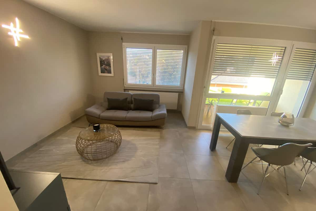 A living room with a sofa, a round coffee table, and a dining table with chairs. There are windows with blinds, a picture on the wall, and a staircase leading to another level.