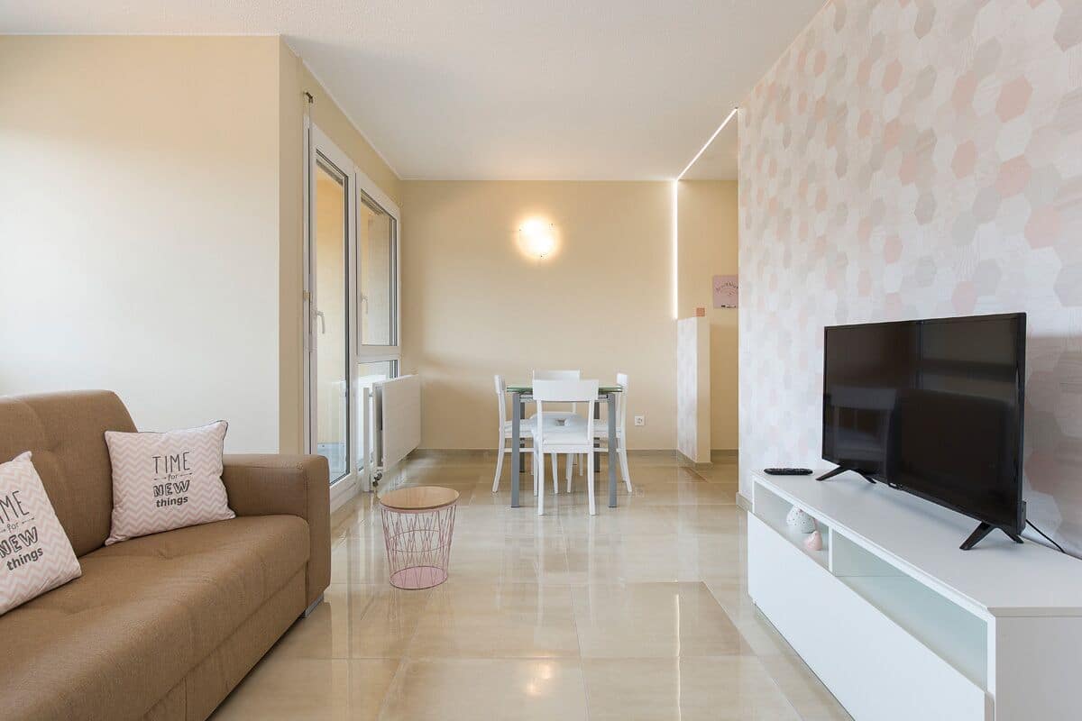 A modern living room with a sofa, a TV on a stand, a dining table with chairs, and a patterned wall.