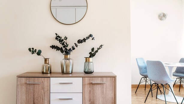A dining area with a wooden cabinet, three vases with plants, a round mirror, and blue chairs around a white table. - Pazzallo Apartments (Lugano)