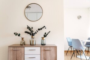 Un comedor con un mueble de madera, tres jarrones con plantas, un espejo redondo y sillas azules alrededor de una mesa blanca.